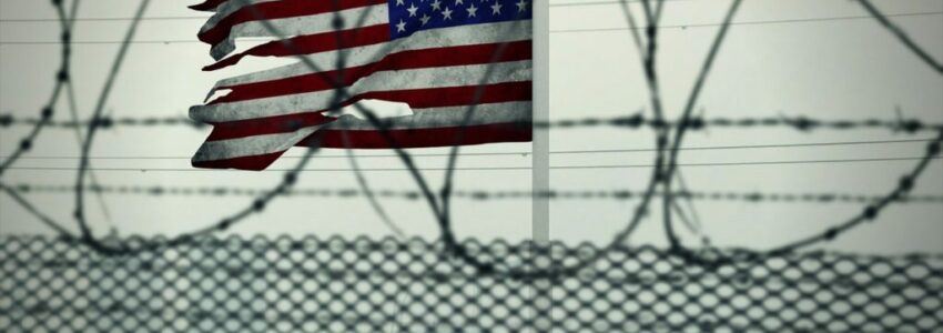 A tattered US Flag flying above a private prison fence.