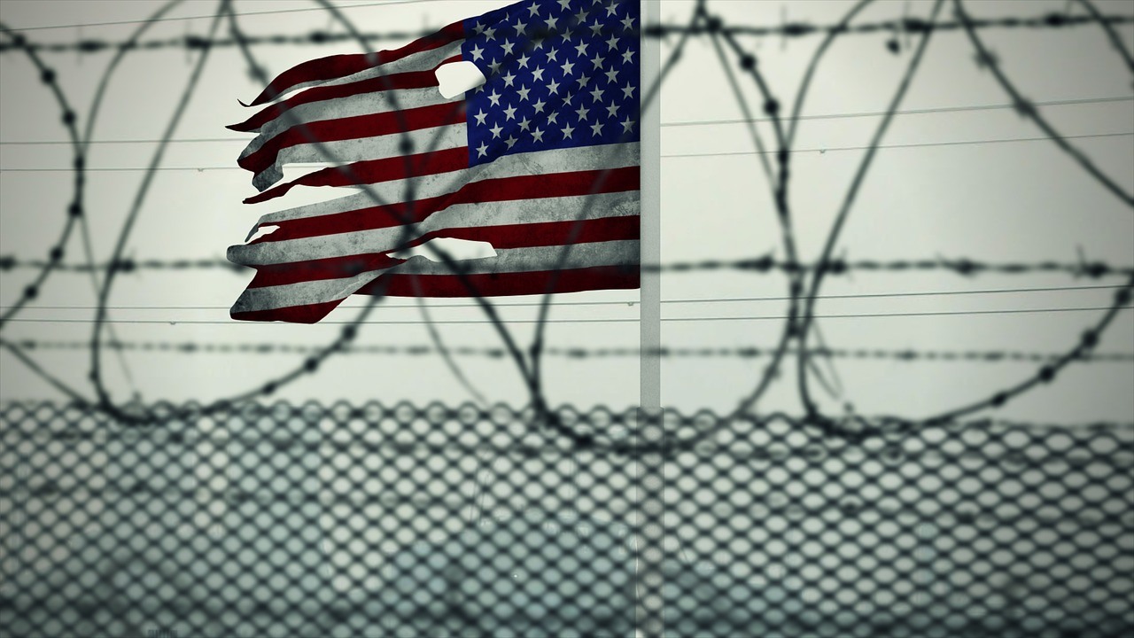 A tattered US Flag flying above a private prison fence.