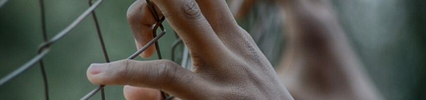 A pair of hands belonging to a young prisoner of color leaning against the prison fence.