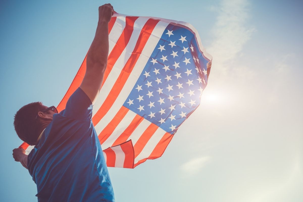 A man holds up an American flag towards the sun.