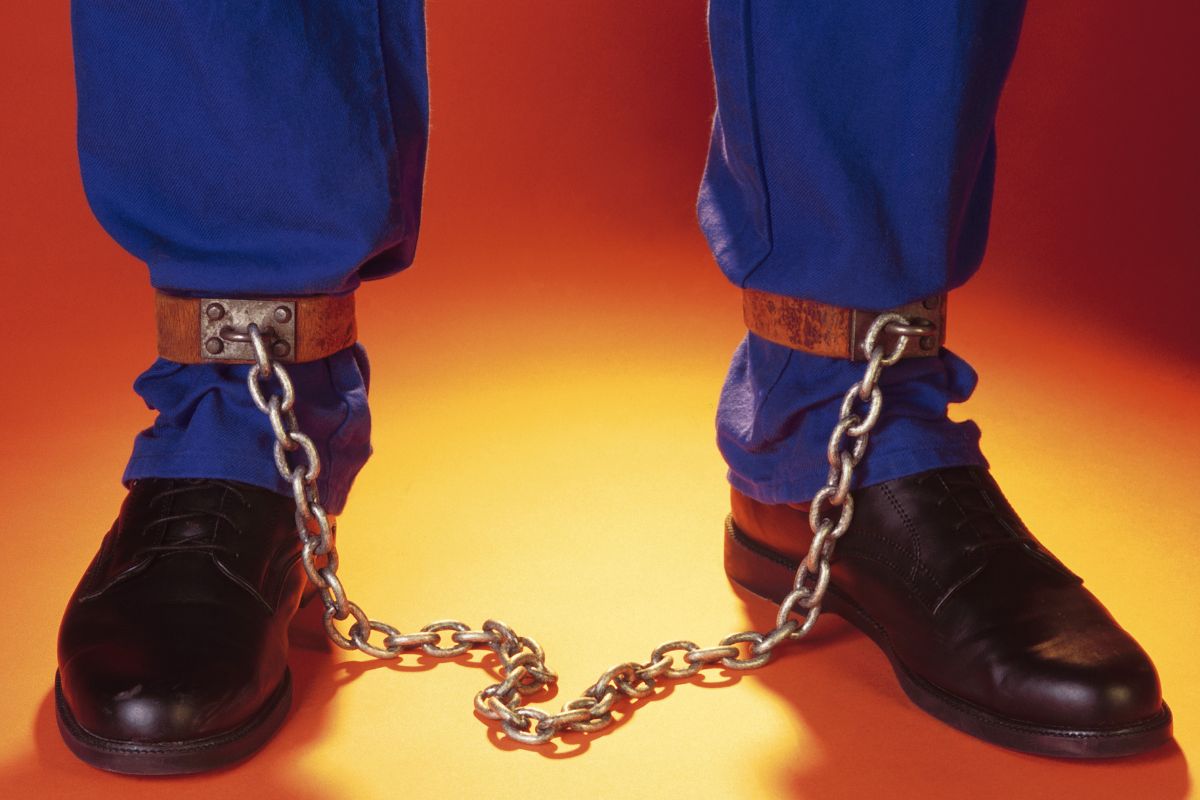 A man's leg with jail shoes and shackles on both ankles.